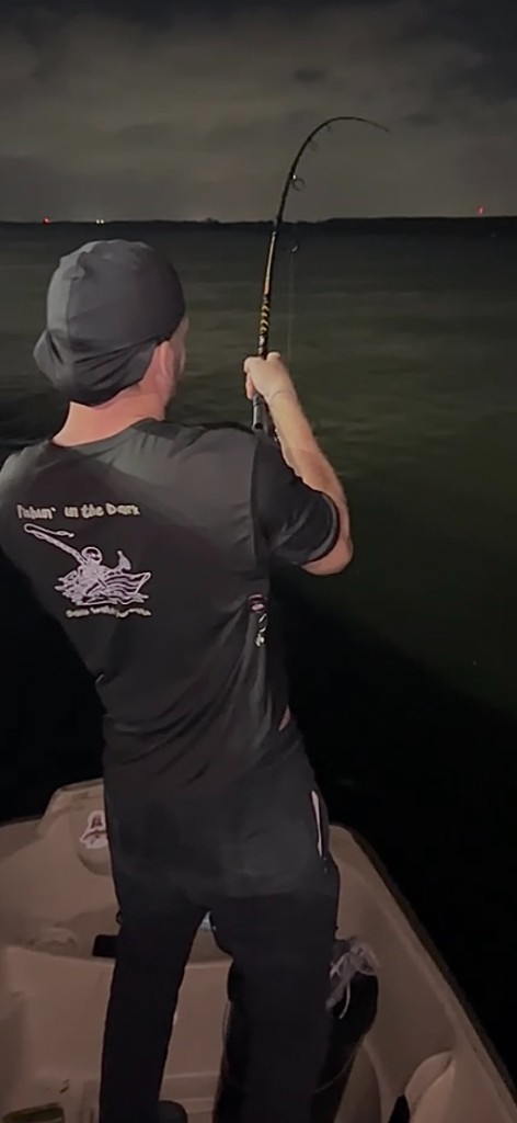 Angler on the water at night wearing a Fishin' in the Dark t-shirt, rod bent on a fish